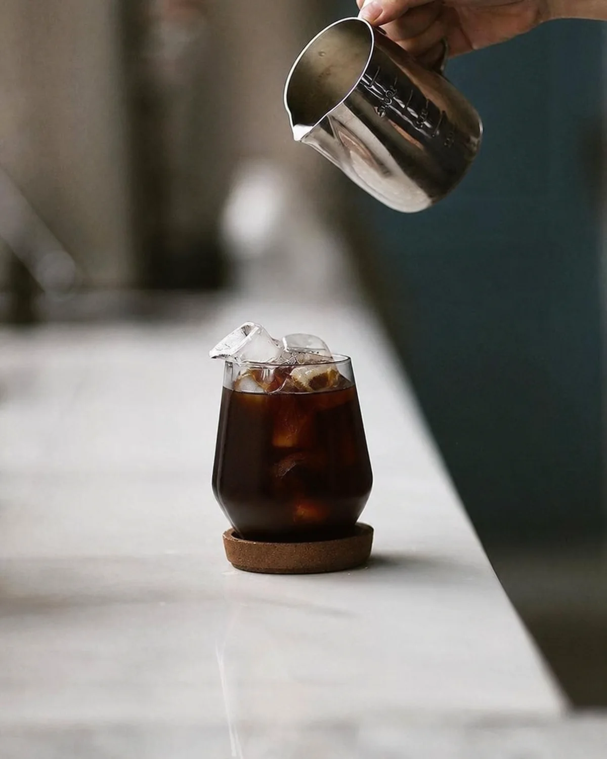 Metal pitcher held above a glass of dark iced coffee